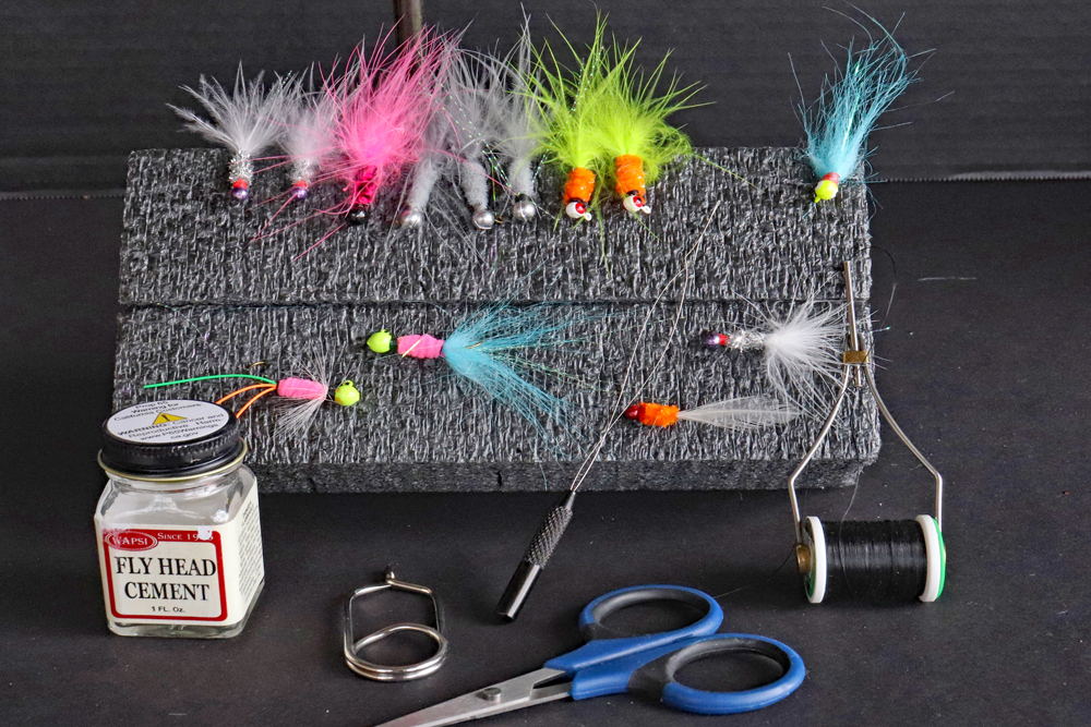 A small sampling of hand-tied crappie jigs, plus the necessary tools for tying. Left to right, the tools are head cement, hackle pliers, bobbin threader, bobbin and scissors.