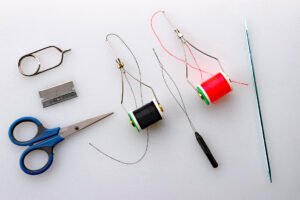 In addition to a good vise, the necessary tools for jig tying include hackle pliers, razor blade, scissors, two bobbins with thread and bobbin threader between them, head cement application tool.